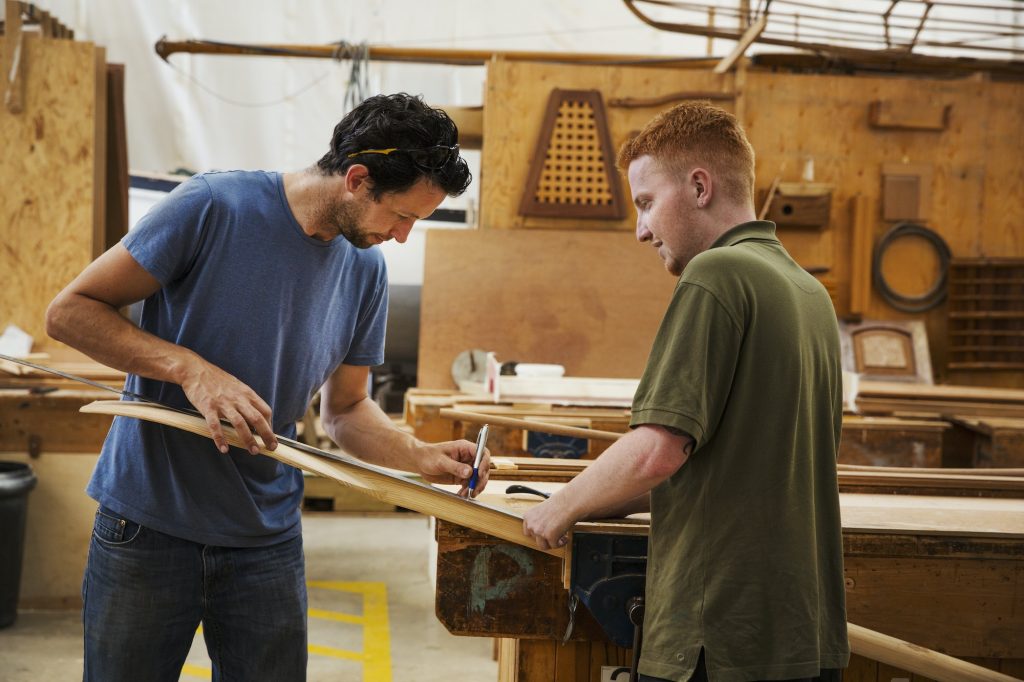 Two men standing at a workbench in a boat-builder's workshop, working on piece of wood.